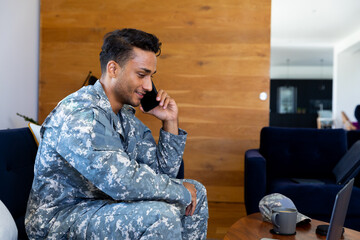 Happy biracial male soldier wearing uniform, sitting on sofa talking on smartphone in living room