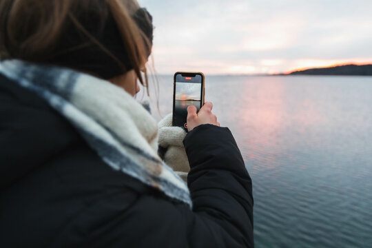 Young woman taking photo with her phone on the ocean in the sunset 