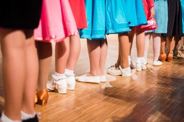 Young girls ballroom dancers standing in a row before beginners competition. Colorful dresses and different shoes on wooden floor. Kids ballroom dancing. - obrazy, fototapety, plakaty