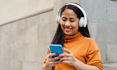 Happy southeast Asian woman listening to music with mobile smartphone and headphones in the city...