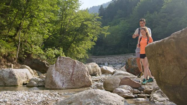 Young Couple, Walking Along The Rocky Shore Of A Mountain River, Exploring Beautiful Nature, Low Angle Shot. Concept Of An Active Vacation.