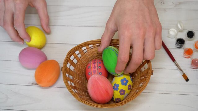 Human hands put colored eggs into basket on white table. Happy Easter concept. Unrecognized caucasian man preparing beautiful holy symbols for religious holiday