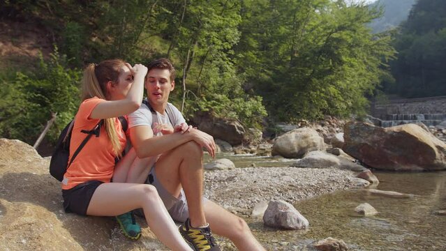 Young couple sitting and relaxing on the mountain river shore, enjoying nature and a sunny summer day, handheld shot. Concept of the romantic weekend and active pastime.