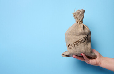 A female hand holds a burlap sack with the inscription subsidy on a blue background, concept of...