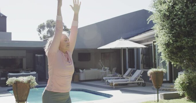 Happy senior caucasian woman doing yoga, stretching by swimming pool in garden, slow motion