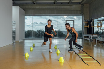 Full length of personal trainer encouraging boy during circuit training routine in gym. Jumping through cones, quick functional exercise in a large gym with windows at night. Horizontal