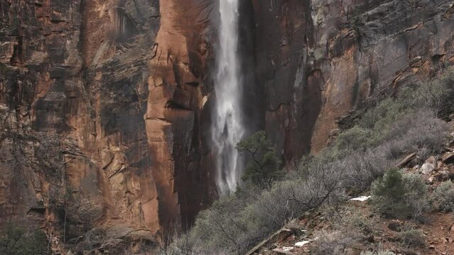 The water from an ephemeral waterfall crashes down a red and black streaked sandstone cliff in slow motion in front of a pine tree near the historic tunnel, after heavy rains in Zion Nat. park, Utah.