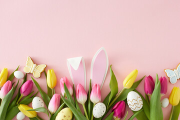 Easter mood idea. Flat lay composition of rabbit bunny ears white yellow easter eggs tulips flowers and butterfly cookies on pastel pink background with copyspace