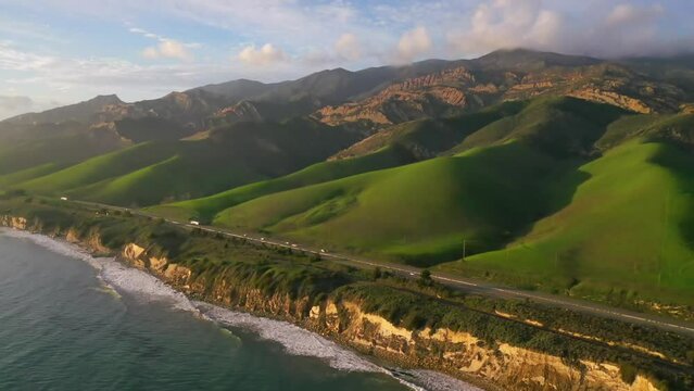 2023 - Excellent aerial footage pulling away from ocean waves lapping the beach on the coast of Gaviota, California while traffic drives by on the 101 highway on a sunny day.