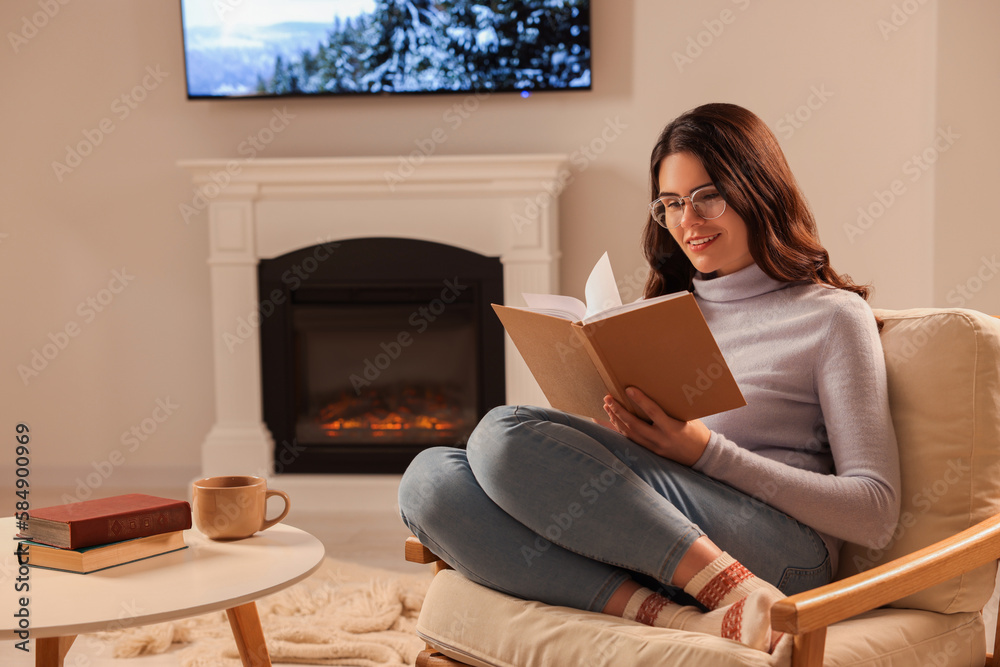 Poster Young woman reading book in armchair near fireplace at home