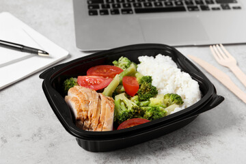 Container with tasty food, laptop, cutlery and notebook on light grey table. Business lunch