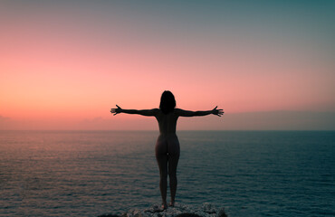 Standing woman with open arms on cliff at sunset