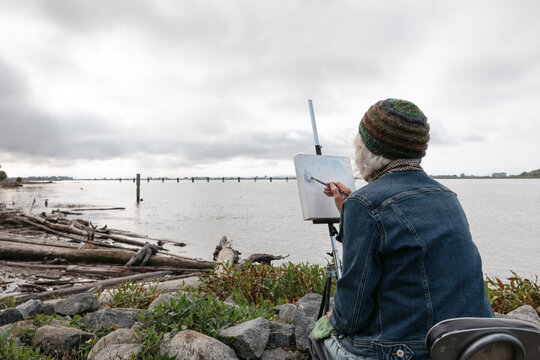 Silver haired woman painting on canvas by the river.