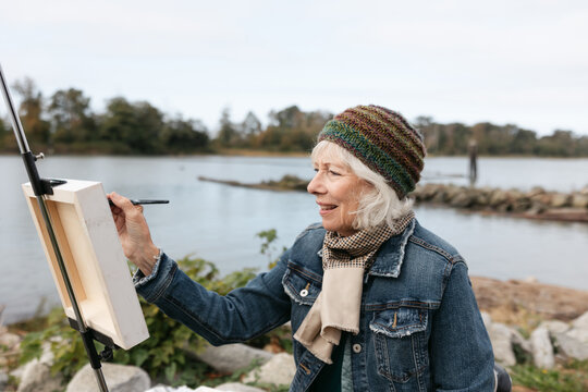 Silver haired woman painting on canvas by the river.