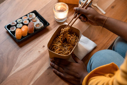 Crop black woman eating ordered noodles and sushi

