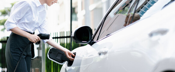 Closeup progressive businesswoman with smartphone, leaning electric car and charging station before...