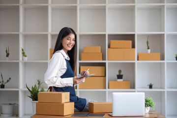 Fototapeta na wymiar A portrait of a young Asian woman, e-commerce employee sitting in the office full of packages in the background write note of orders and a calculator, for SME business ecommerce and delivery business