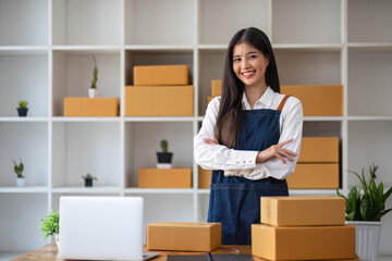 Fototapeta na wymiar A portrait of a young Asian woman, e-commerce employee sitting in the office full of packages in the background write note of orders and a calculator, for SME business ecommerce and delivery business