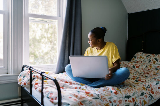 Smiling black girl sitting on her bed in her teenage room on laptop 