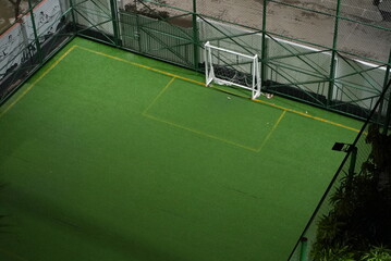 A beautiful, green, empty football playground with a white-colored goalpost, surrounded by green trees, green fence made of net and a street on the background. 