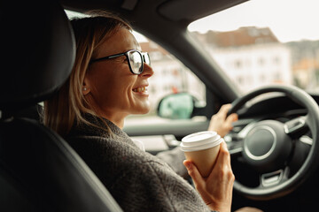 Smiling businesswoman wearing eyeglasses sitting behind steering wheel in car and drinking coffee 