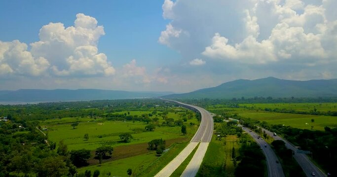 High angle view of the capturing a curved and parallel road path between the green rice fields at sunlight with beautiful clouds and blue sky.