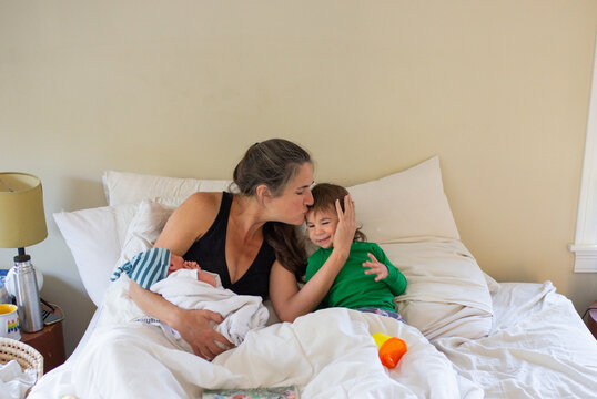 Postpartum Mother Kisses her Toddler While Holding New Baby in Bed