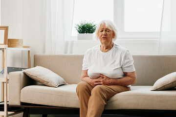 Fototapeta na wymiar Elderly woman severe abdominal pain sitting on the sofa, health problems in old age, poor quality of life. Grandmother with gray hair holding her stomach, poisoning, problems with stool.