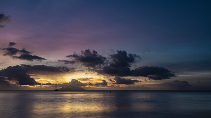 Fantastically beautiful tropical sunset. Purple clouds in the sky, highlighted in pink and gold. Reflection on the calm surface of the ocean. Silhouette of an island and a yacht on the horizon. 