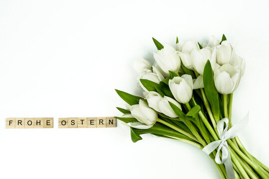 Blumenstrauß aus weißen Tulpen mit weißer Schleife rechts im Bild,  weißeR Hintergrund, links daneben mit hölzernen Spielsteinen die Worte FROHE OSTERN gelegt, links oben noch freier Platz, horizontal