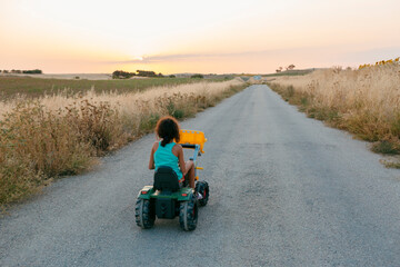 Kid riding a toy tractor on countryside road at sunset in a summer day