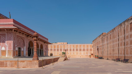 The courtyard of the ancient City Palace is tiled. The walls of the building are built of red sandstone, decorated with white ornaments. Arches, colonnade, lighting lanterns are visible. Blue sky. 