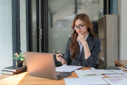 Image of a young beautiful brooding Asian woman working with a laptop while sitting at a laptop in the office, thinking of a professional plan, project management, and considering new business ideas.