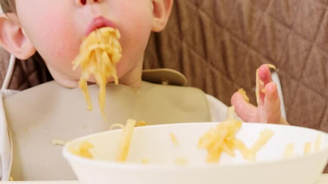 A Funny Child Is Eating A Grated Apple With His Mouth Full While Sitting On A Kitchen Chair. Hungry Baby Boy Shoves Food In His Mouth, Humor. Kid Aged One Year And Three Months