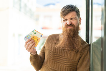 Redhead man with beard taking a lot of money at outdoors with sad expression