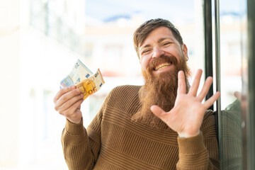Redhead man with beard taking a lot of money at outdoors saluting with hand with happy expression