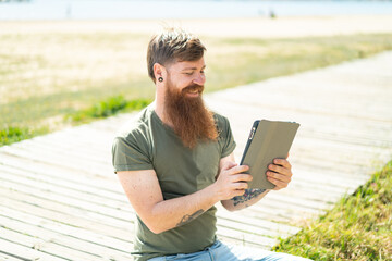 Redhead man with beard holding a tablet at outdoors with happy expression