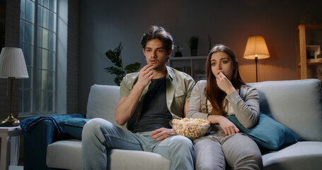 Young student caucasian concentrated couple sitting on couch in the evening watching tv show and...