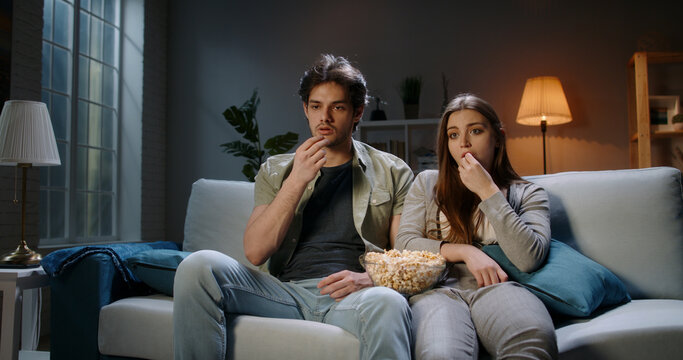 Young student caucasian concentrated couple sitting on couch in the evening watching tv show and eating popcorn. Millennials watching their favourite series after hard day