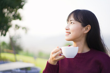 Happy beautiful Asian young woman sitting on the grass field and enjoy relaxing with a stunning view of mountain and forest landscape. Asian young woman drinking a cup of coffee in morning.