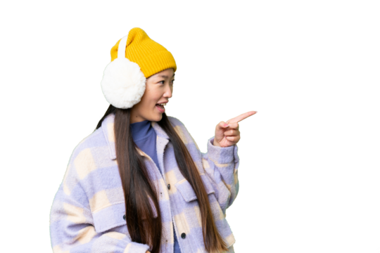 Young Asian woman wearing winter muffs over isolated chroma key background pointing finger to the side and presenting a product