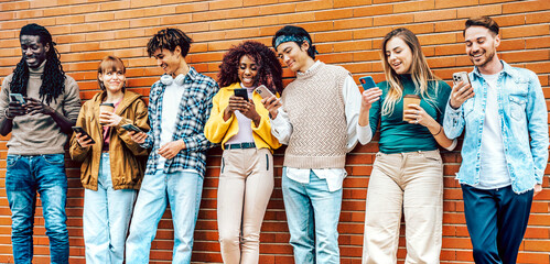 Group of happy friends laugh and joke while using smart mobile phones - Group of multiracial students leaning on a wall using cellular standing outdoors - Concept of youth, tech, social and friendship