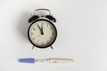 Flat lay with black retro alarm clock and pregnancy test kit showing positive result, isolated white backdrop. Pregnancy, motherhood and gynecological concept. Women's health and fertility. Still life