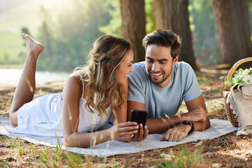 Couple on picnic with smartphone, relax together in nature and social media with travel and bonding outdoor. Happiness, man and woman lying down and scroll internet in forest with smile