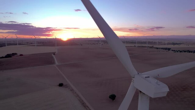 Aerial view of a wind turbine farm with a close-up of a wind turbine spinning as seen from the nacelle at sunset. A group of windmills are turning in the background. Cuenca, Castilla La Mancha, Spain