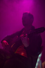 Musician playing acoustic guitar in a foggy club with colorful lights.