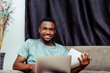 A man sits on a couch with a laptop and a black curtain behind him.