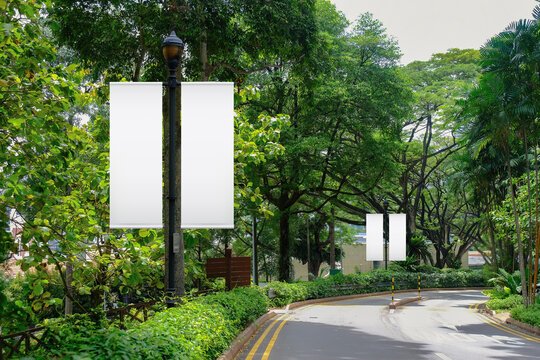 Blank vertical advertising banners on street lampposts; double hanging posters by the road, against lush green trees and plants. For OOH out of home template mock up