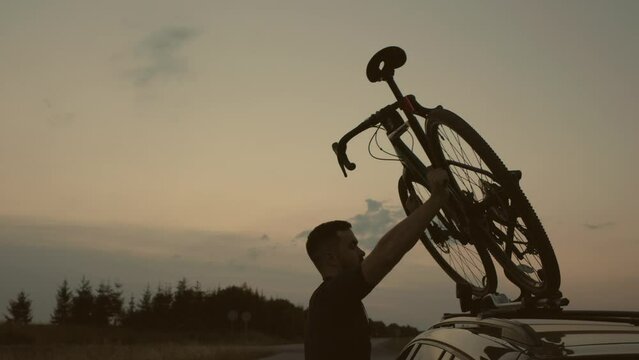 A man mounts his bicycle on the roof of a car at sunset. The concept of outdoor recreation.
