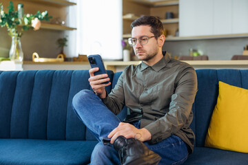 Man Using Smartphone At Home. Handsome Guy Sitting On Sofa And Messaging In Cozy Living Room at Home. Male Person Doing Online Shopping, Browsing Internet and Checking Social Media